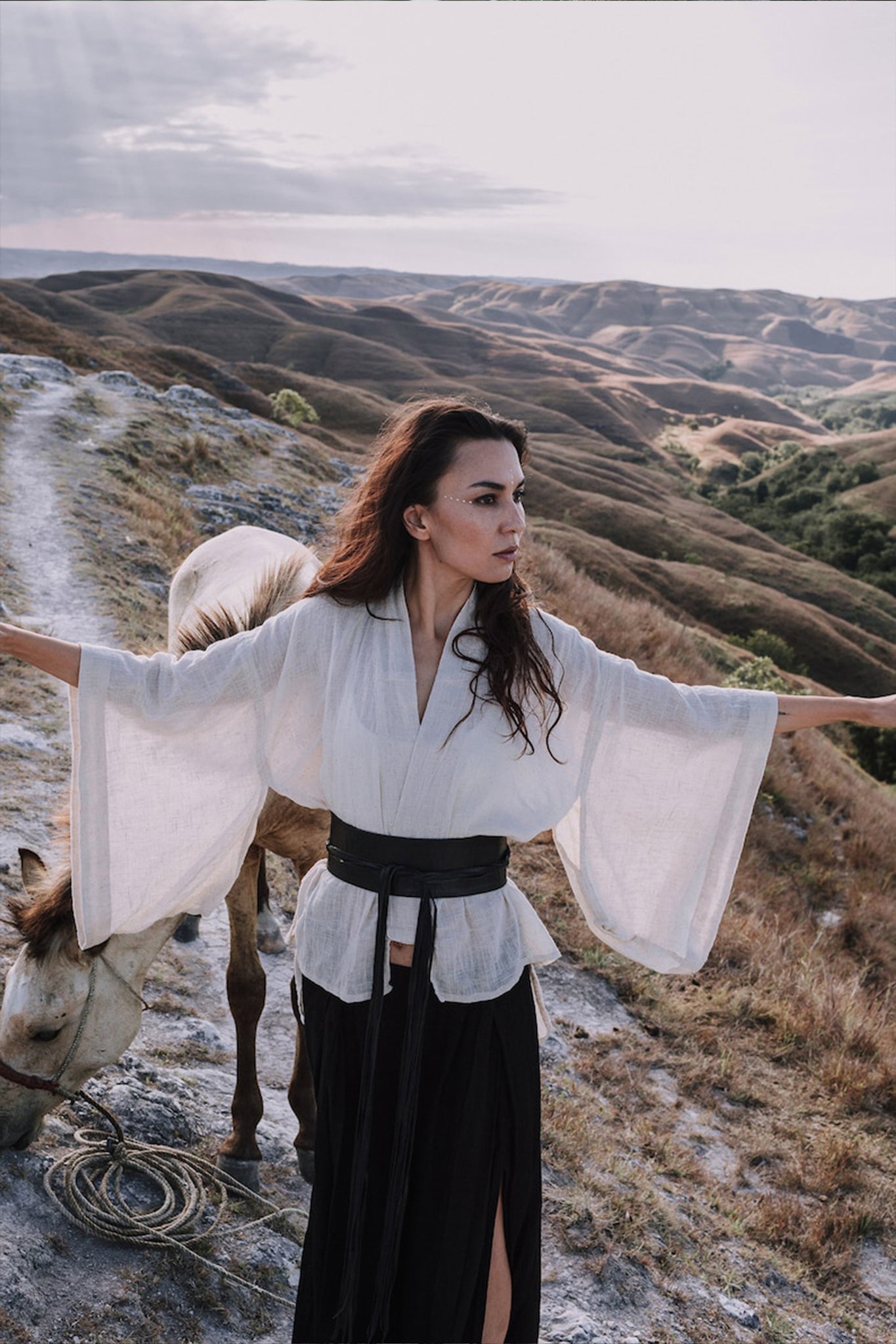 A woman stands on a scenic hillside with outstretched arms, wearing an elegant Off-White Kimono Top from AYA Sacred Wear and a black skirt. A horse is tied nearby. Rolling hills and a cloudy sky form the backdrop, underscoring her connection to sustainable production.