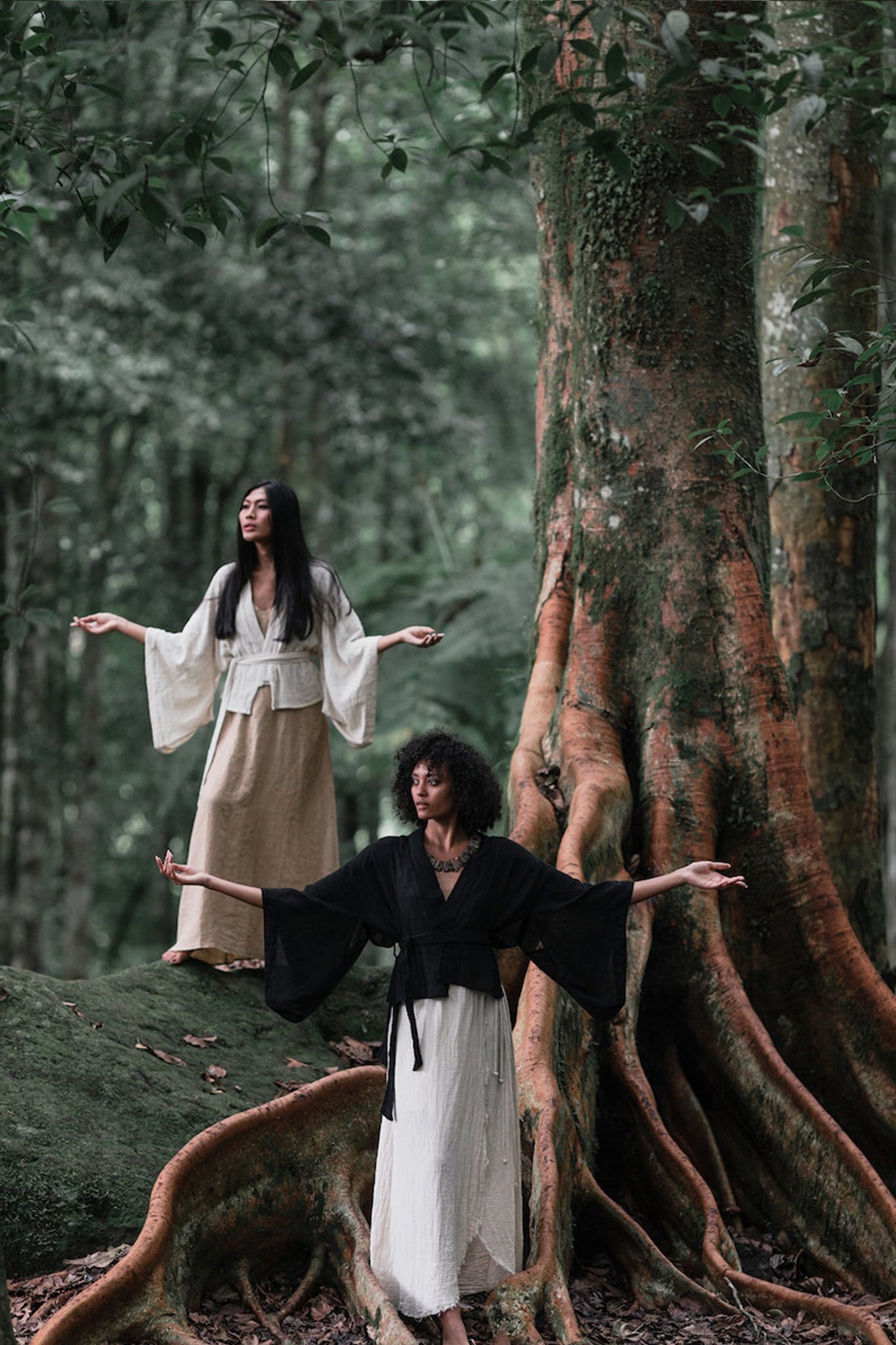 Two individuals stand on tree roots in a forest, their arms outstretched. They wear flowing outfits—one in organic cotton white and beige, the other featuring the Off-White Kimono Top from AYA Sacred Wear. A large tree trunk and lush greenery surround them, creating a serene and sustainable setting.