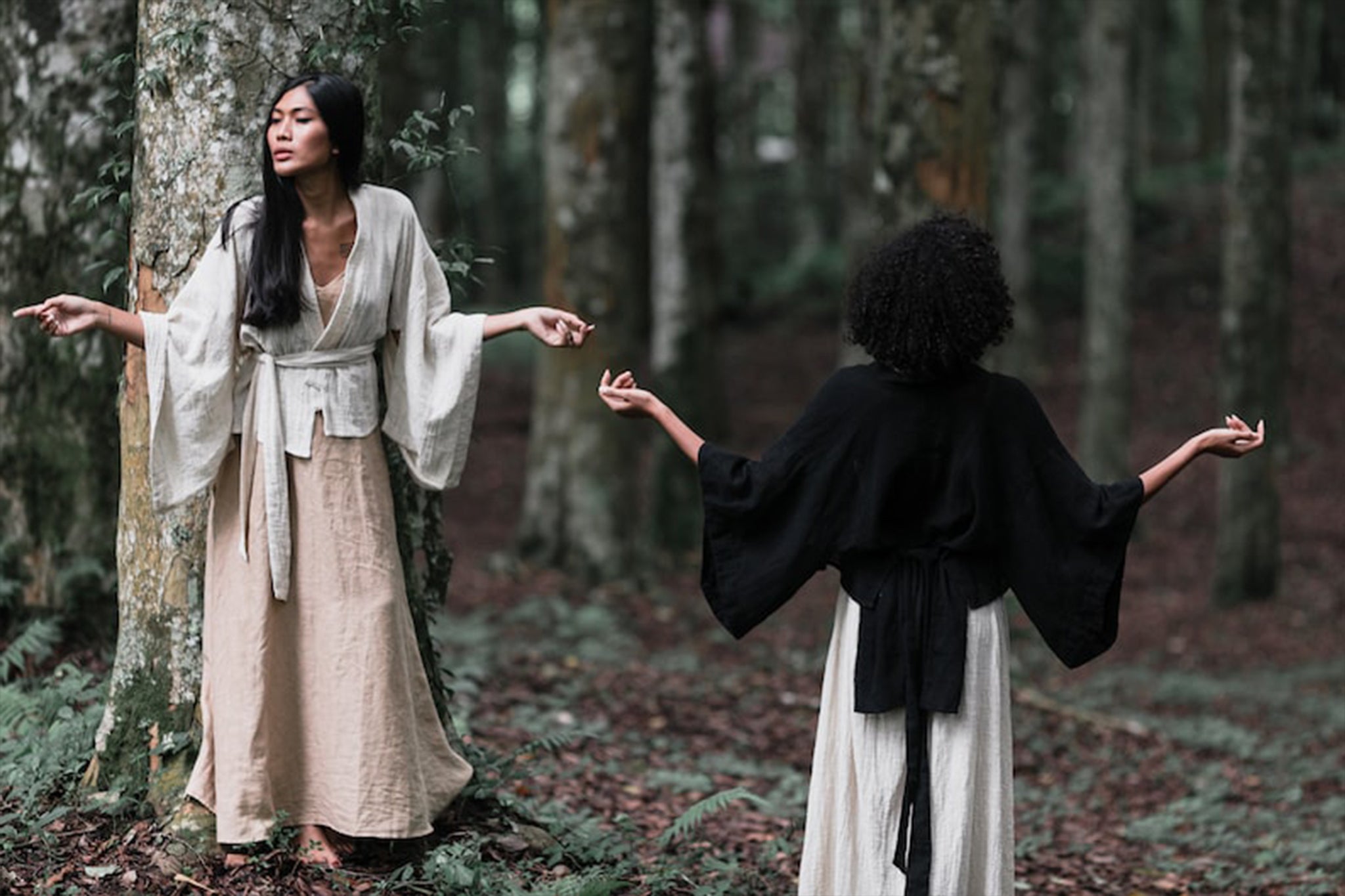 Two people dressed in flowing, loose garments stand amidst a forest backdrop. One leans against a tree, looking at the camera, while the other faces away with their arms gently outstretched. They are showcasing the Off-White Kimono Top • Tribal Boho Top from AYA Sacred Wear, made of organic cotton and surrounded by towering trees and lush greenery.