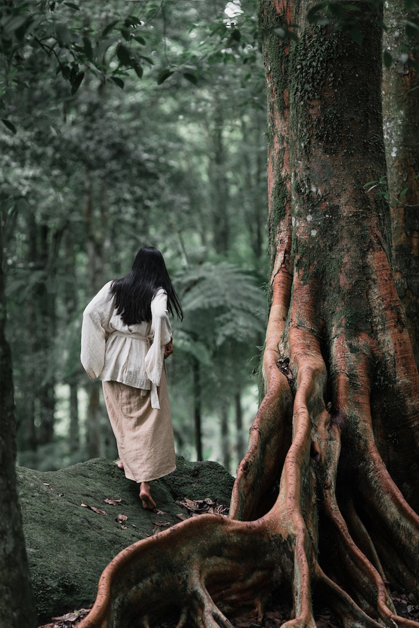 A person with long, dark hair strolls barefoot through a lush forest, wearing an Off-White Kimono Top from AYA Sacred Wear paired with a beige skirt. They are near a large tree with exposed roots, surrounded by dense greenery.