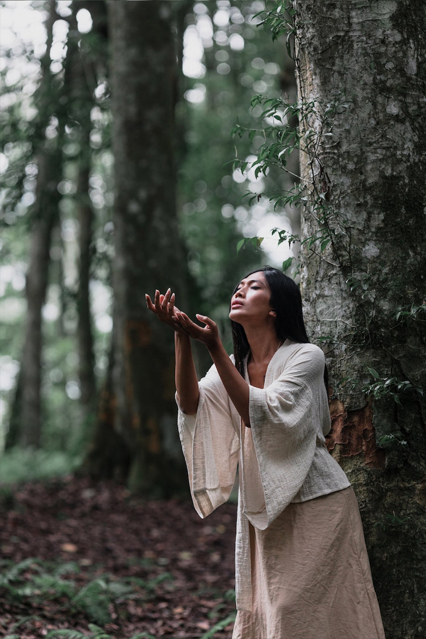 Dressed in an Off-White Kimono Top by AYA Sacred Wear, a woman stands gracefully in a forest, clad in a beige skirt. Her organic cotton ensemble harmonizes with the calm environment as she leans against a tree, eyes closed and hands raised, fully immersed in the vibrant greenery around her.