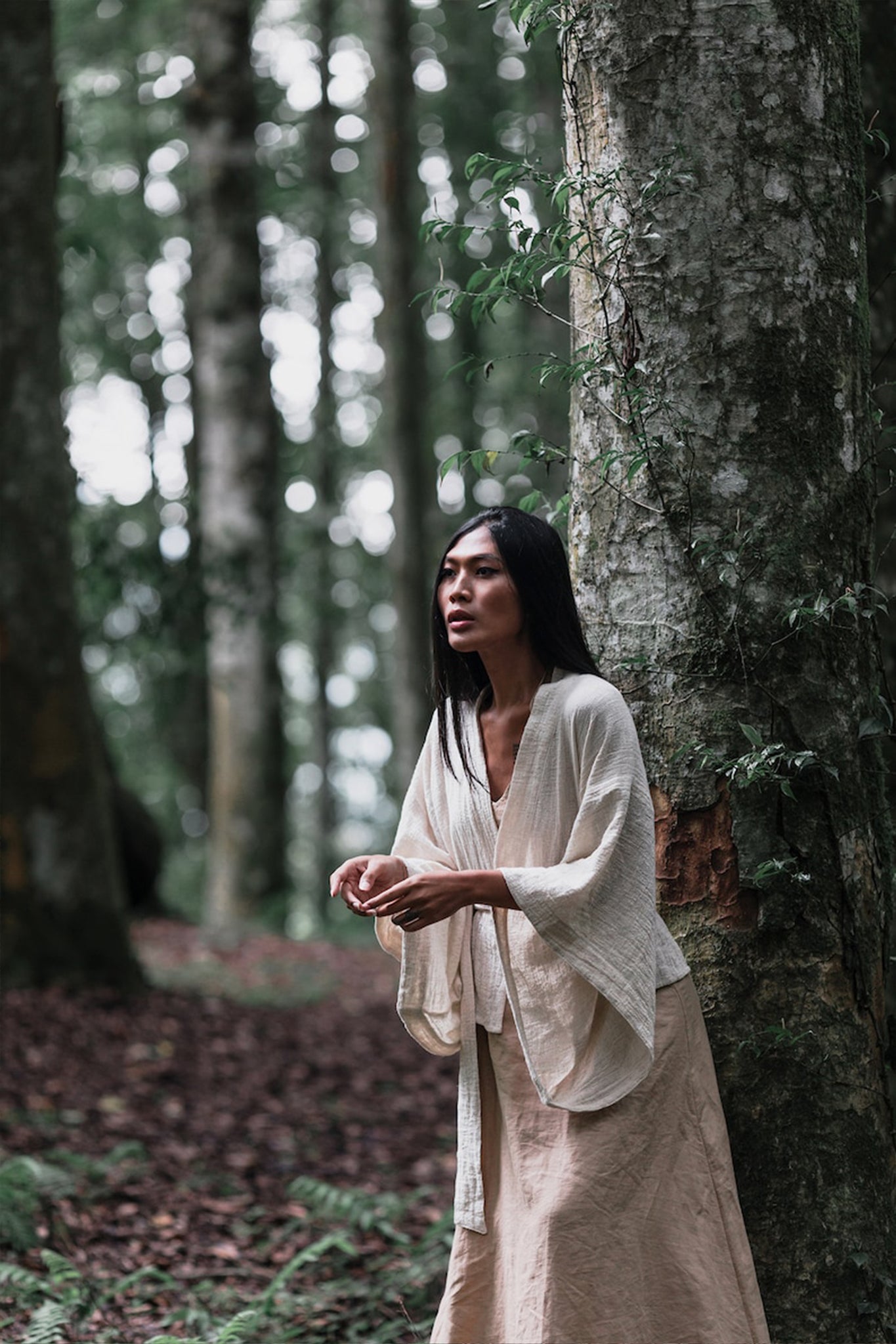 A woman with long black hair, wearing the Off-White Kimono Top from AYA Sacred Wear, stands by a tree in a dense forest. She appears contemplative, surrounded by tall trees and lush greenery, her Tribal Boho Top reflecting sustainable production with organic cotton woven into its fabric.