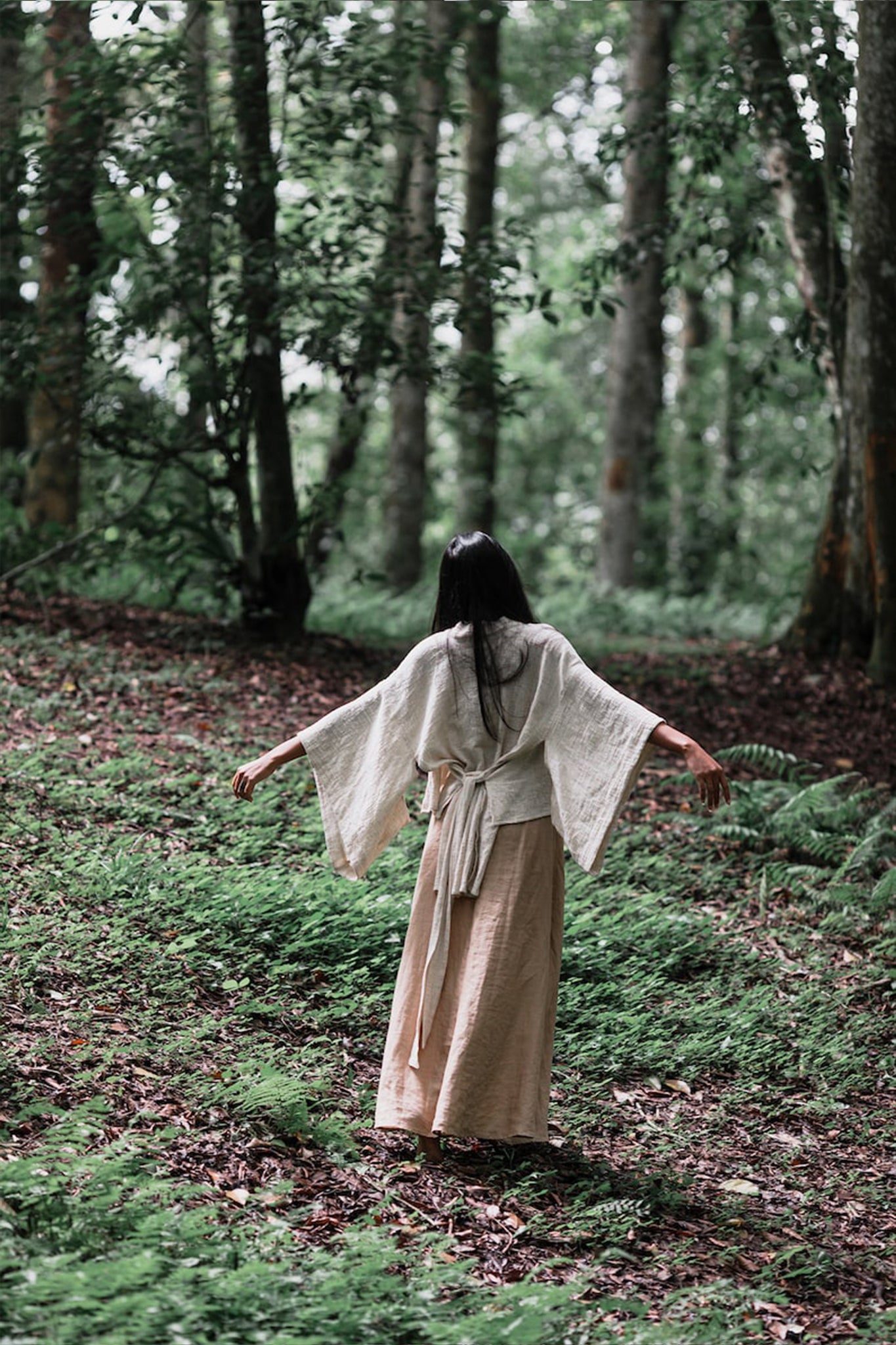 Wearing the Off-White Kimono Top by AYA Sacred Wear, a person stands with arms outstretched in a serene forest, surrounded by tall trees and lush greenery. Made from organic cotton through sustainable production, this Tribal Boho Top enhances the scene's sense of peace and connection with nature.