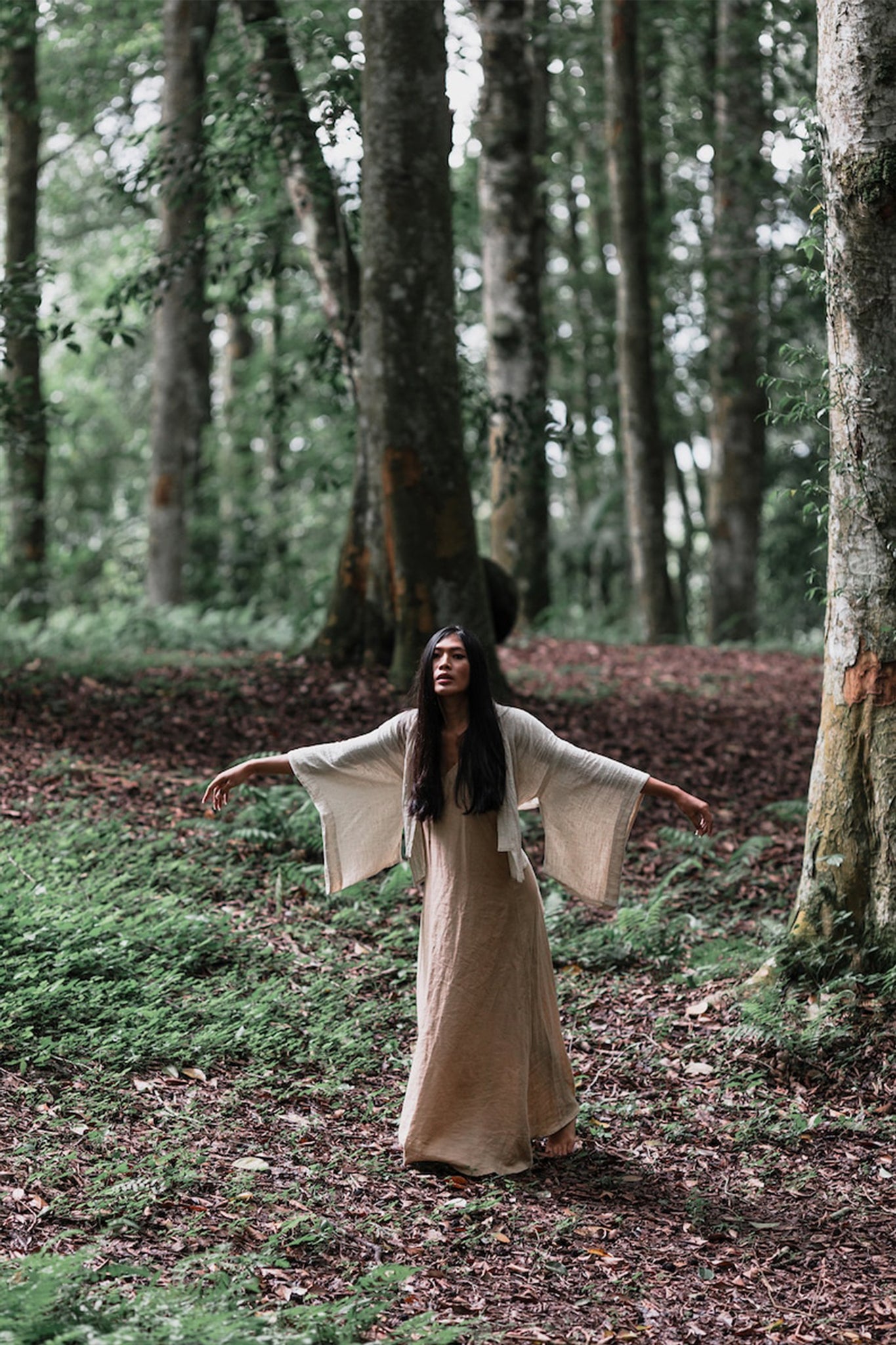 A woman dressed in an Off-White Kimono Top from AYA Sacred Wear, designed in a Tribal Boho style, stands with her arms outstretched in the heart of a lush forest, where tall trees stretch towards the sky and vibrant green foliage carpets the earth beneath scattered leaves and ferns.