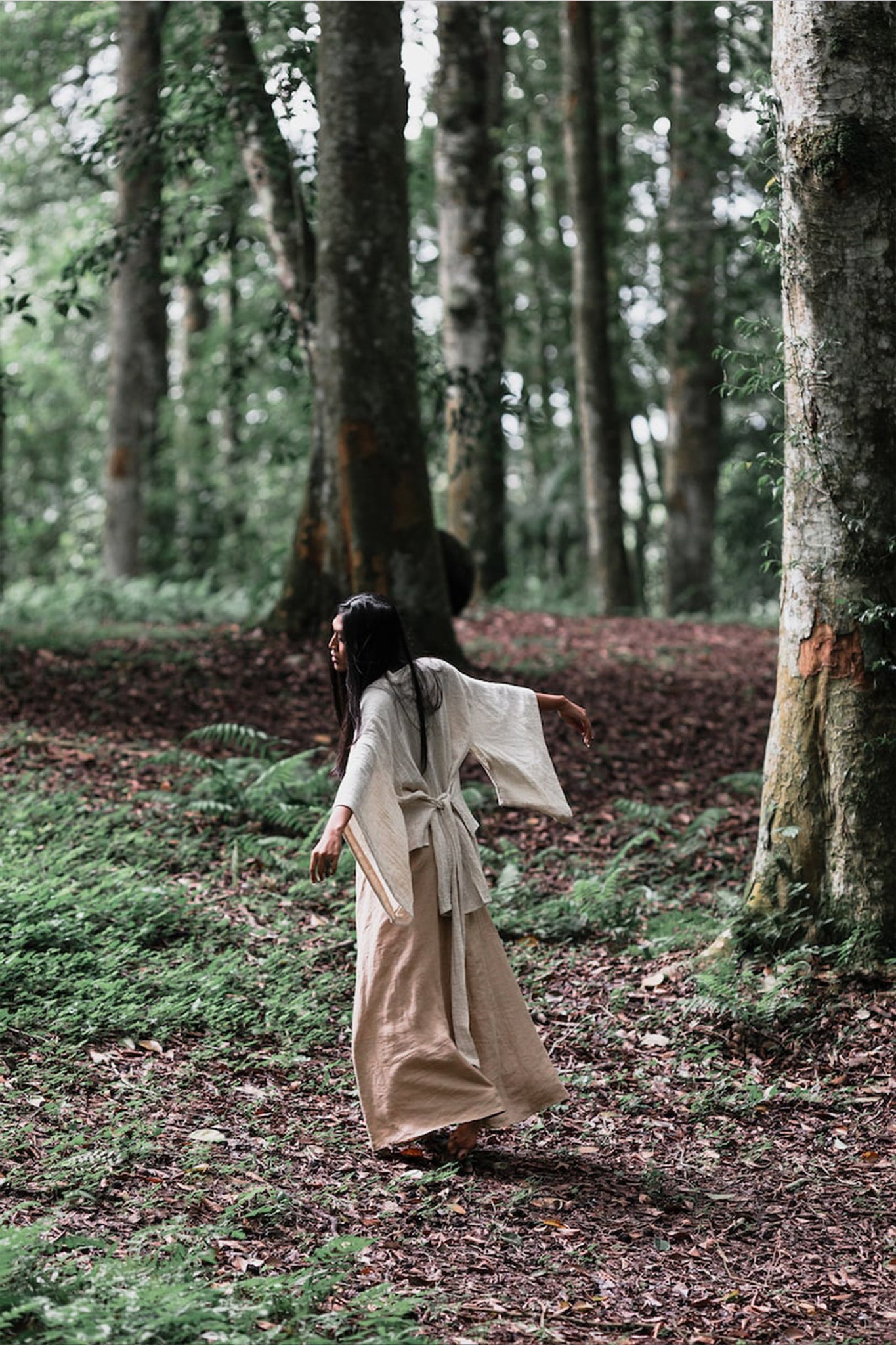 Dressed in an off-white kimono top from AYA Sacred Wear, a person meanders through a verdant forest, enveloped by towering trees and rich greenery, fostering a sense of peace and unity with nature. The Tribal Boho Top embodies sustainable production practices, enhancing the serene experience amidst nature.