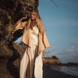 A woman with long hair stands barefoot on a rocky beach, wearing an ensemble featuring the Off-White Linen Boho Pants with High Slit by AYA Sacred Wear. Her contemplative gaze meets the clear sky, dotted with clouds, as the ocean stretches in the background. This scene of tranquility embodies the spirit of sustainable fashion.