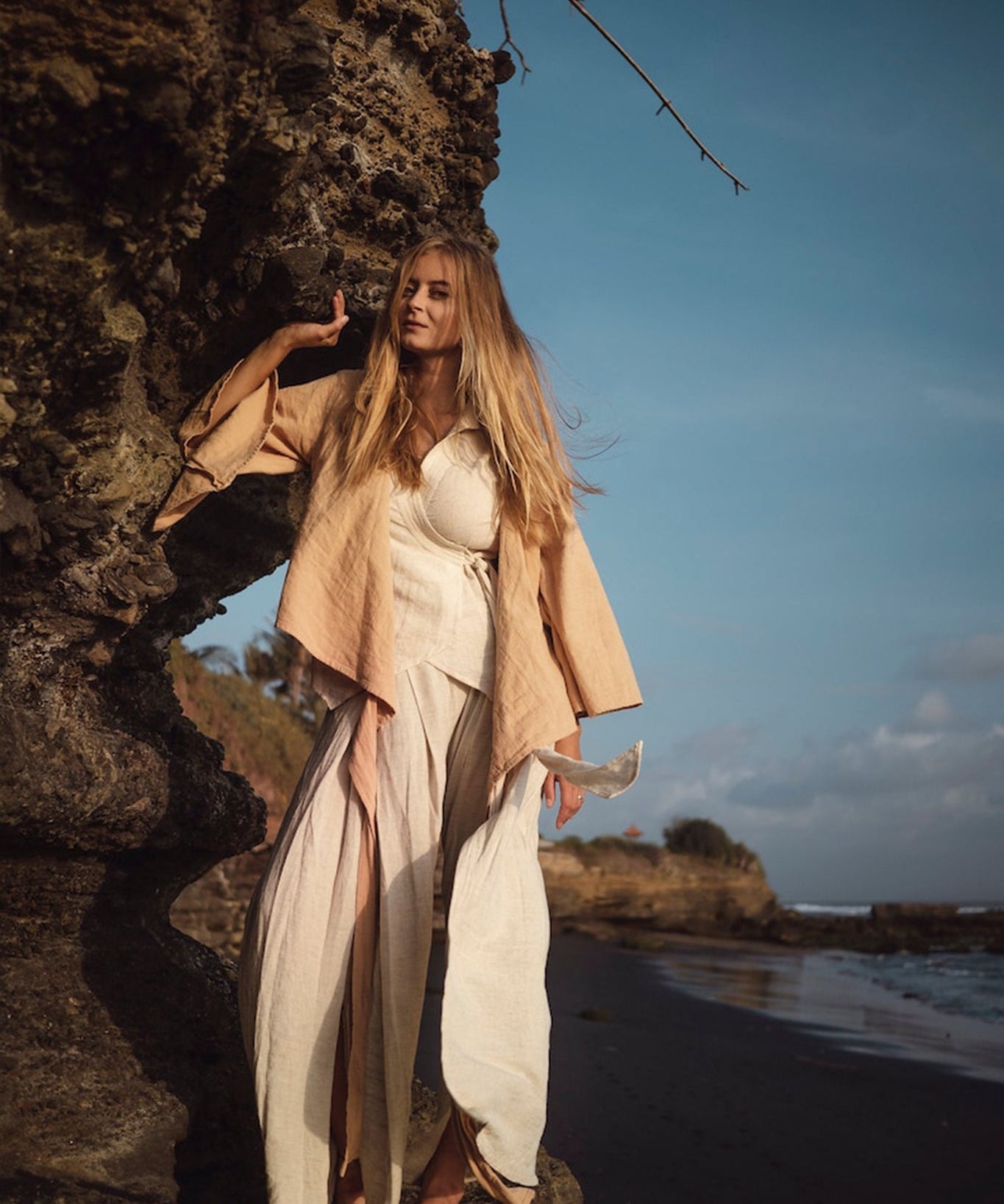 A woman with long hair stands barefoot on a rocky beach, wearing an ensemble featuring the Off-White Linen Boho Pants with High Slit by AYA Sacred Wear. Her contemplative gaze meets the clear sky, dotted with clouds, as the ocean stretches in the background. This scene of tranquility embodies the spirit of sustainable fashion.