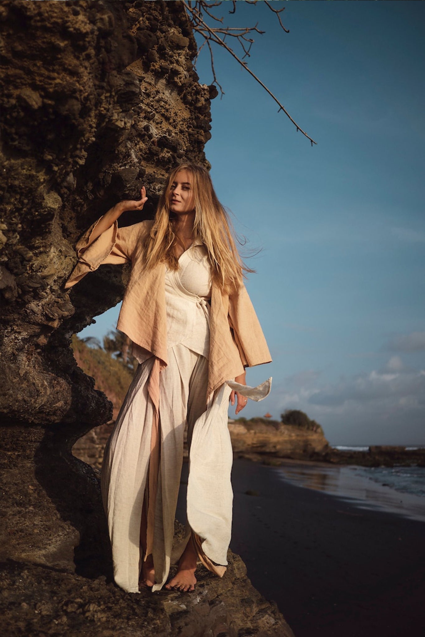 A woman with long hair stands barefoot on a rocky beach, wearing an ensemble featuring the Off-White Linen Boho Pants with High Slit by AYA Sacred Wear. Her contemplative gaze meets the clear sky, dotted with clouds, as the ocean stretches in the background. This scene of tranquility embodies the spirit of sustainable fashion.