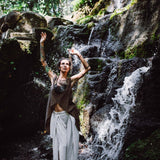 Standing barefoot beside a cascading waterfall, a person wears Off-White Linen Boho Pants with High Slit from AYA Sacred Wear along with a sleeveless top. Their arms are lifted upwards amidst lush greenery and sunlight streaming through the trees.