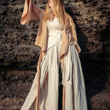 Standing against a rugged stone wall, a person with long hair showcases the essence of sustainable fashion by wearing AYA Sacred Wear's Off-White Linen Boho Pants with High Slit. With one arm raised, the natural texture of the rock provides a striking contrast to the airy elegance of these wide-legged trousers.