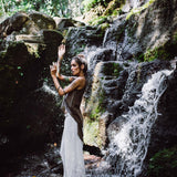 Amidst a lush, green forest beside a small waterfall, a woman poses elegantly in off-white linen boho pants with a high slit by AYA Sacred Wear. The sunlight filtering through the trees highlights her commitment to sustainable fashion as it illuminates the water and surrounding foliage.