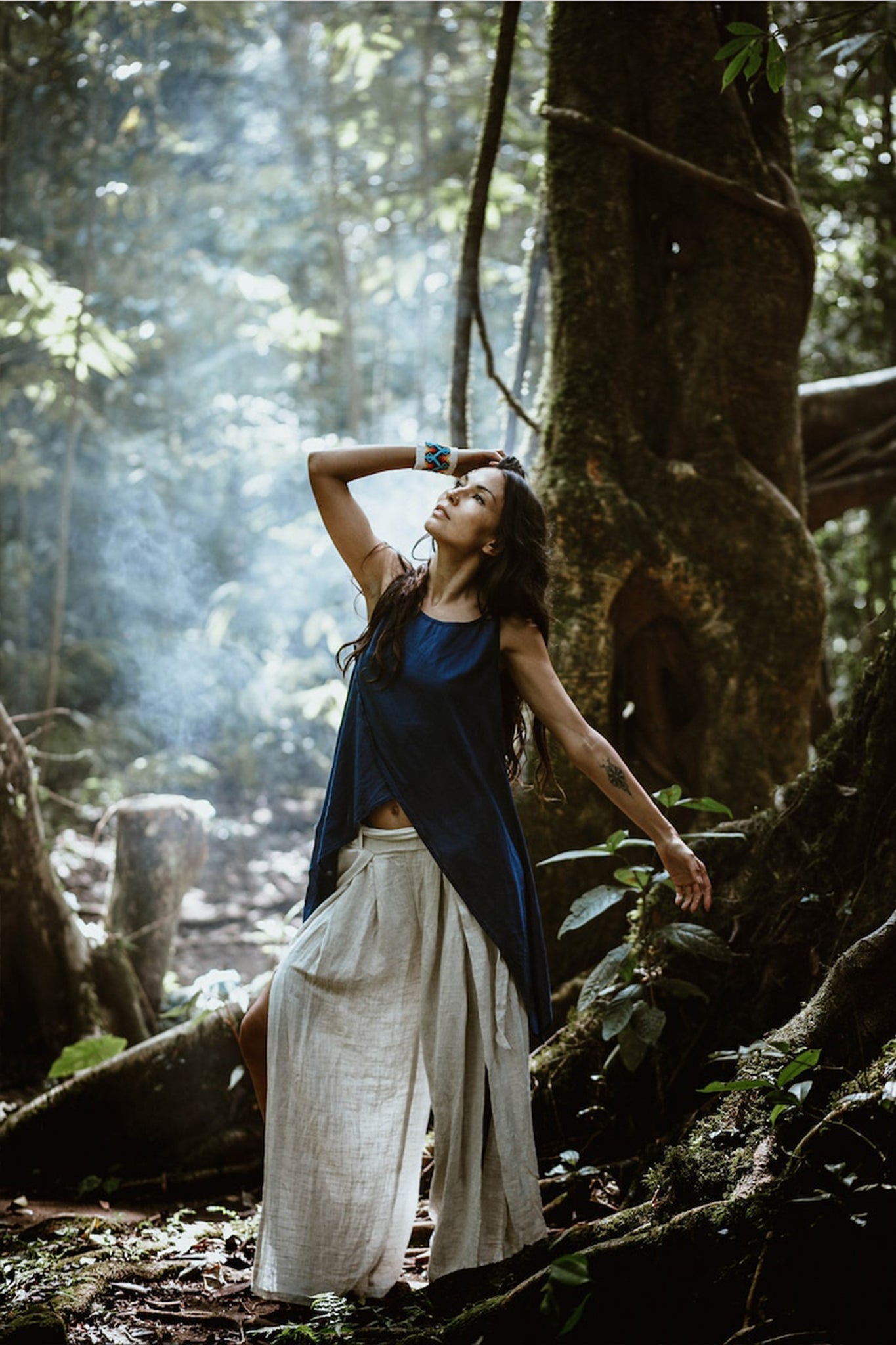 A woman in a blue sleeveless top and Off-White Linen Boho Pants with High Slit from AYA Sacred Wear poses thoughtfully among tall trees in a sunlit forest, with beams of light filtering through the canopy. She has long dark hair and wears a colorful bracelet. The scene is serene and natural.