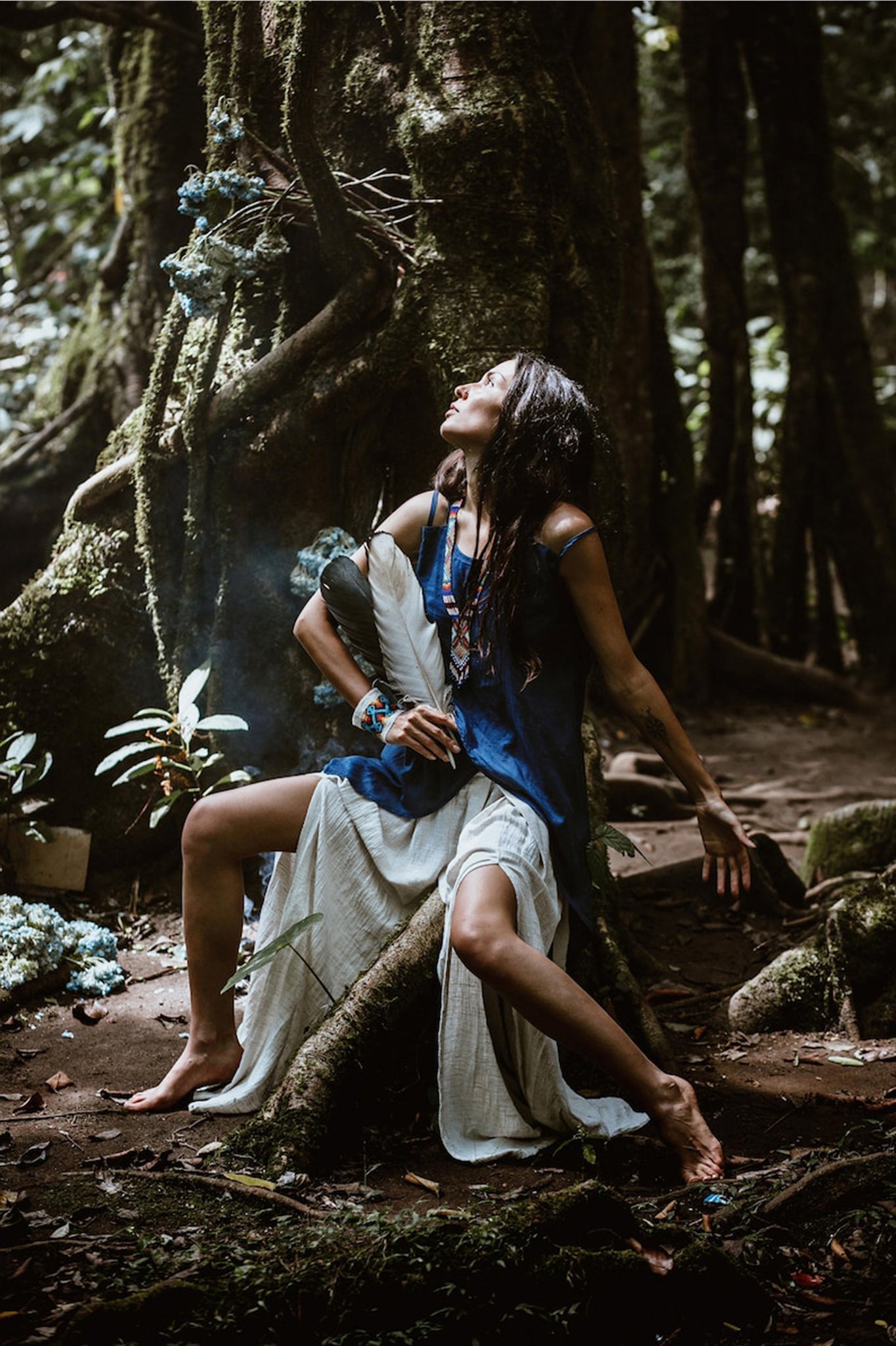 A woman wearing Off-White Linen Boho Pants with a High Slit from AYA Sacred Wear sits on a tree root in the forest, holding a large feather. Surrounded by lush greenery and flowers, with smoke rising nearby, she appears relaxed and contemplative. Her serene expression complements the natural beauty of her surroundings, reminiscent of sustainable production's harmony.