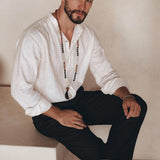 A man with short dark hair and a beard sits on a step indoors, wearing the Off-White Linen Classic Minimalist Shirt by AYA Sacred Wear. He pairs it with dark pants and sandals, while a long beaded necklace drapes loosely around his neck. The organic linen of his shirt perfectly complements his look as he gazes towards the camera with a neutral expression.