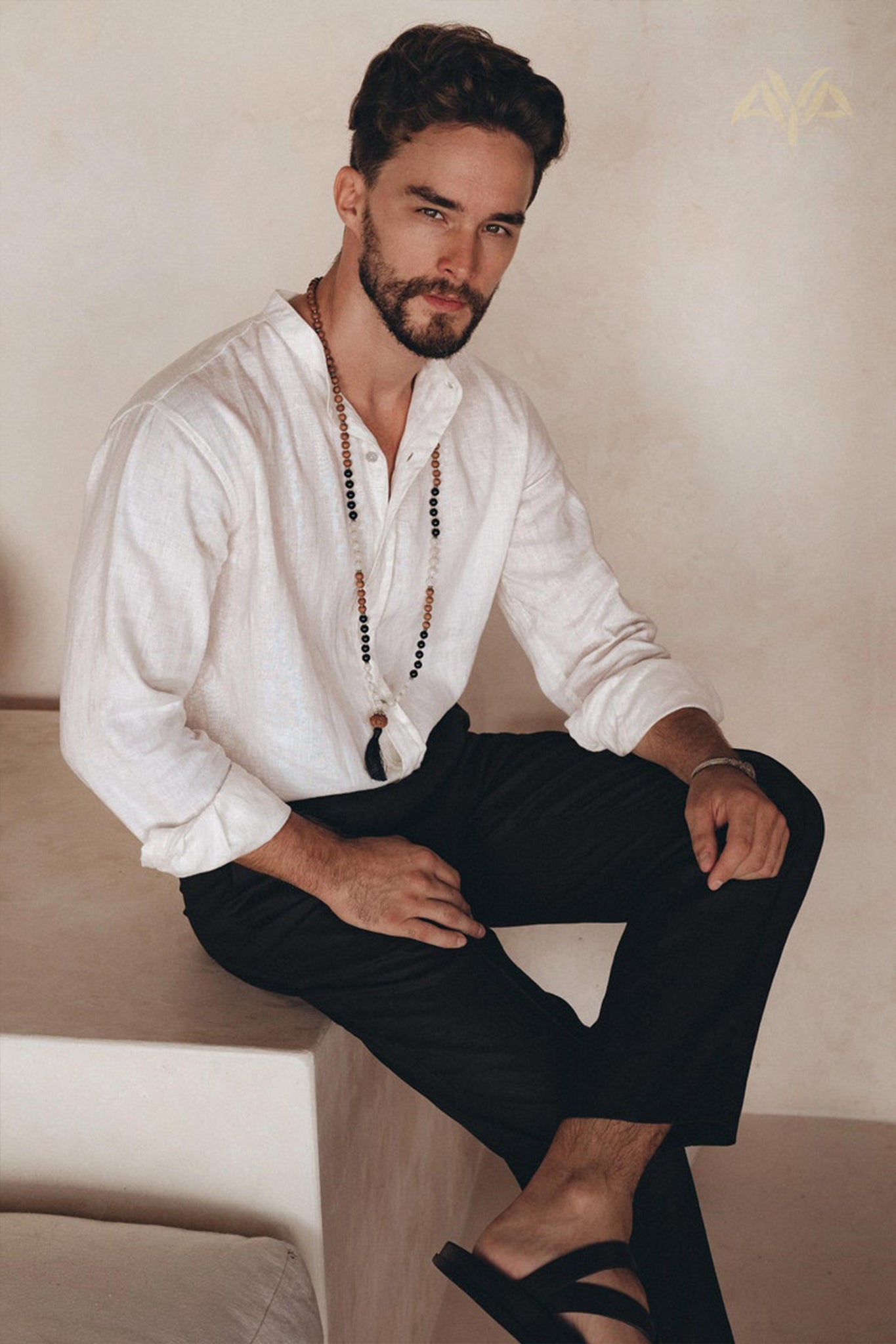 A man with short dark hair and a beard sits on a step indoors, wearing the Off-White Linen Classic Minimalist Shirt by AYA Sacred Wear. He pairs it with dark pants and sandals, while a long beaded necklace drapes loosely around his neck. The organic linen of his shirt perfectly complements his look as he gazes towards the camera with a neutral expression.