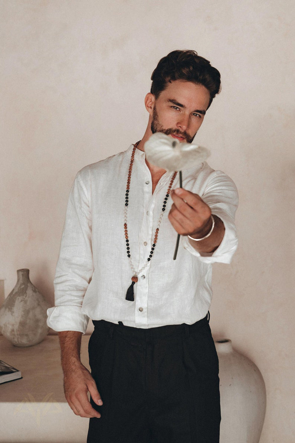 A man wearing an AYA Sacred Wear Off-White Linen Classic Minimalist Shirt and black pants holds a white flower in focus towards the camera. He accessorizes with a necklace and poses indoors against a neutral background, which features a large vase.