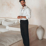 A man with short, dark hair and a beard stands indoors. He is wearing an Off-White Linen Classic Minimalist Shirt for Men from AYA Sacred Wear, black pants, sandals, and a necklace. He poses beside a cream-colored sectional sofa and a large white vase in a minimalist room, embodying the essence of the botanically dyed collection.