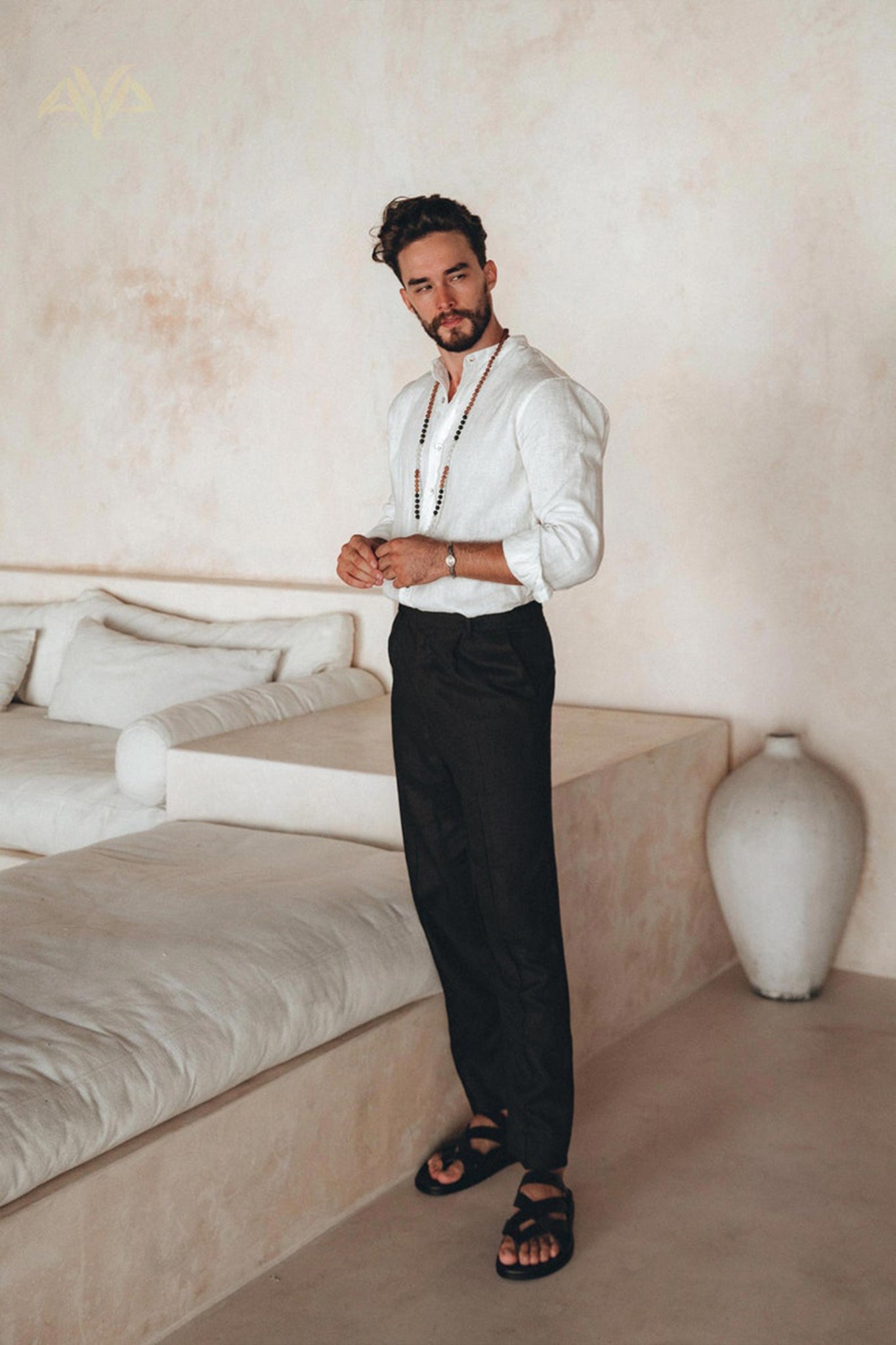 A man with short, dark hair and a beard stands indoors. He is wearing an Off-White Linen Classic Minimalist Shirt for Men from AYA Sacred Wear, black pants, sandals, and a necklace. He poses beside a cream-colored sectional sofa and a large white vase in a minimalist room, embodying the essence of the botanically dyed collection.