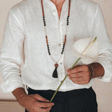 A person wearing an Off-White Linen Classic Minimalist Shirt from AYA Sacred Wear and black pants holds a single white anthurium flower. They have a beaded necklace with a tassel against a neutral background, crafting a serene and elegant scene.