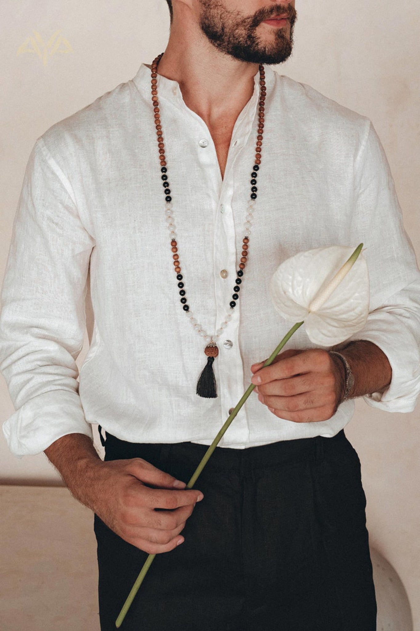 A person wearing an Off-White Linen Classic Minimalist Shirt from AYA Sacred Wear and black pants holds a single white anthurium flower. They have a beaded necklace with a tassel against a neutral background, crafting a serene and elegant scene.