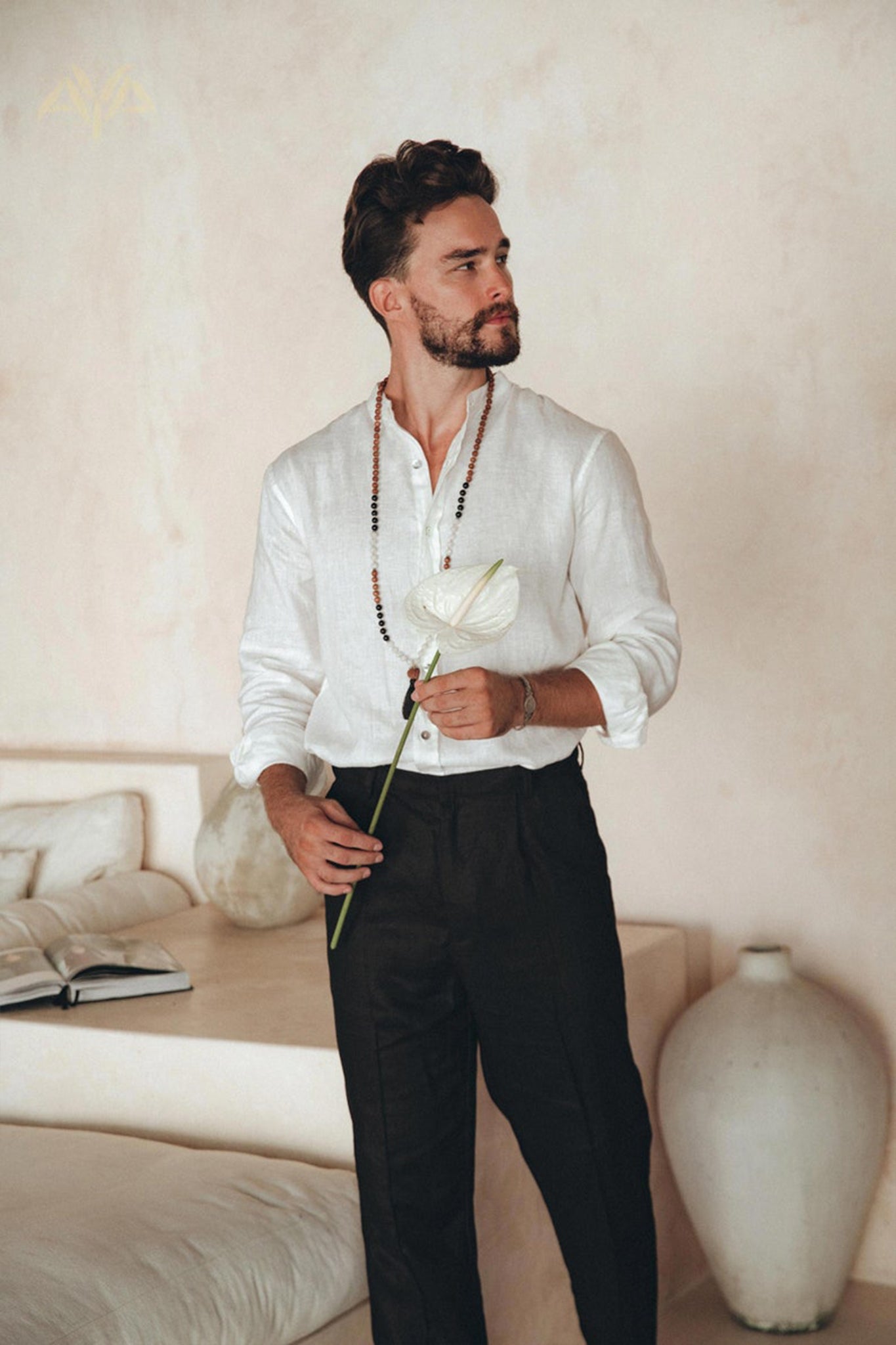 A bearded man in an Off-White Linen Classic Minimalist Shirt by AYA Sacred Wear and black pants stands in a minimalist room, holding a white flower. A beige couch and an open book are visible in the background.