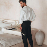 A man with a beard stands on a minimalist, light beige floor next to a white bed, holding a botanically dyed flower. He is dressed in the Off-White Linen Classic Minimalist Shirt by AYA Sacred Wear, paired with black pants and sandals. An open book rests on the bed, and a large vase is placed nearby on the floor.