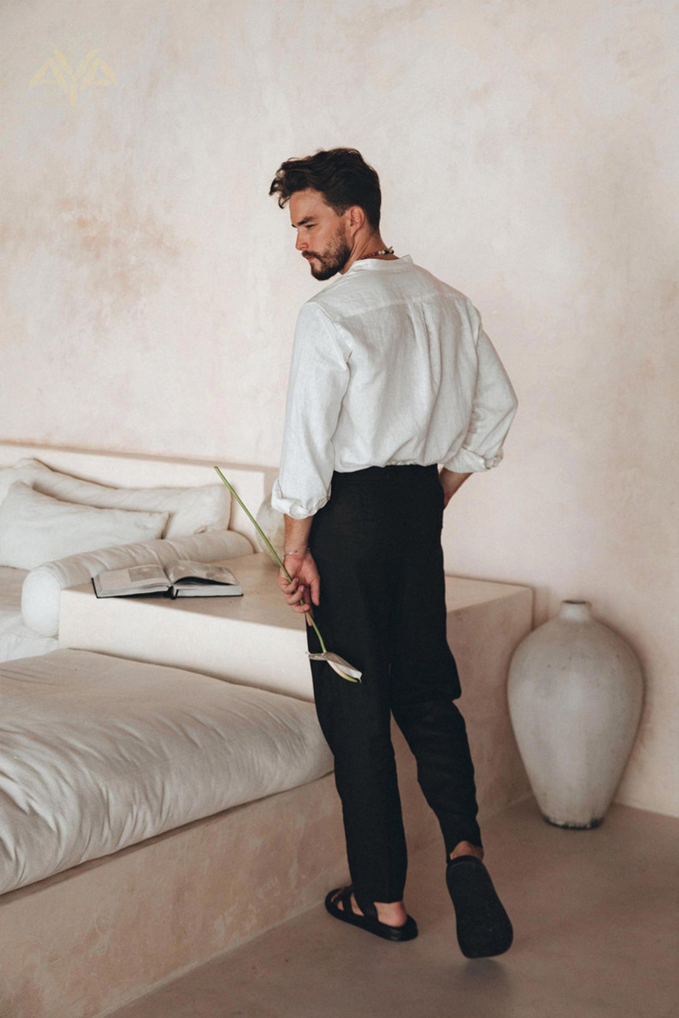 A man with a beard stands on a minimalist, light beige floor next to a white bed, holding a botanically dyed flower. He is dressed in the Off-White Linen Classic Minimalist Shirt by AYA Sacred Wear, paired with black pants and sandals. An open book rests on the bed, and a large vase is placed nearby on the floor.