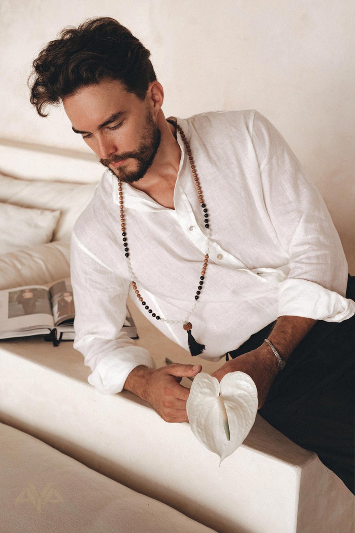A man with dark hair and a beard casually leans on a white couch, wearing the Off-White Linen Classic Minimalist Shirt for Men by AYA Sacred Wear and a beaded necklace. He's holding a white anthurium flower, with an open book beside him, contributing to the serene minimalist atmosphere.