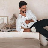 A man in an Off-White Linen Classic Minimalist Shirt from AYA Sacred Wear is lounging on a minimalist, beige sofa, holding a hat. An open book and a decorative vase are beside him, creating a calm and relaxed setting.