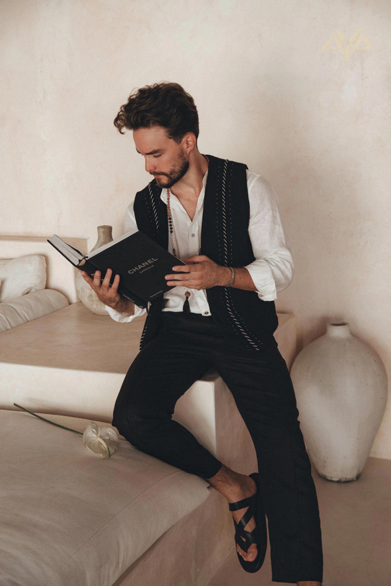 A man with curly hair is sitting on a bed, engrossed in a book. Dressed in an off-white linen classic minimalist shirt from AYA Sacred Wear, paired with a black vest and pants, and sandals on his feet, he exudes effortless style. Beside him lies a white rose from the botanically dyed collection, while a large vase decorates the background.