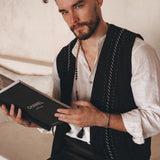 A bearded man wearing an Off-White Linen Classic Minimalist Shirt from AYA Sacred Wear and a black vest sits while holding a Chanel book. He gazes thoughtfully at the camera, with the soft, neutral background creating a warm atmosphere.
