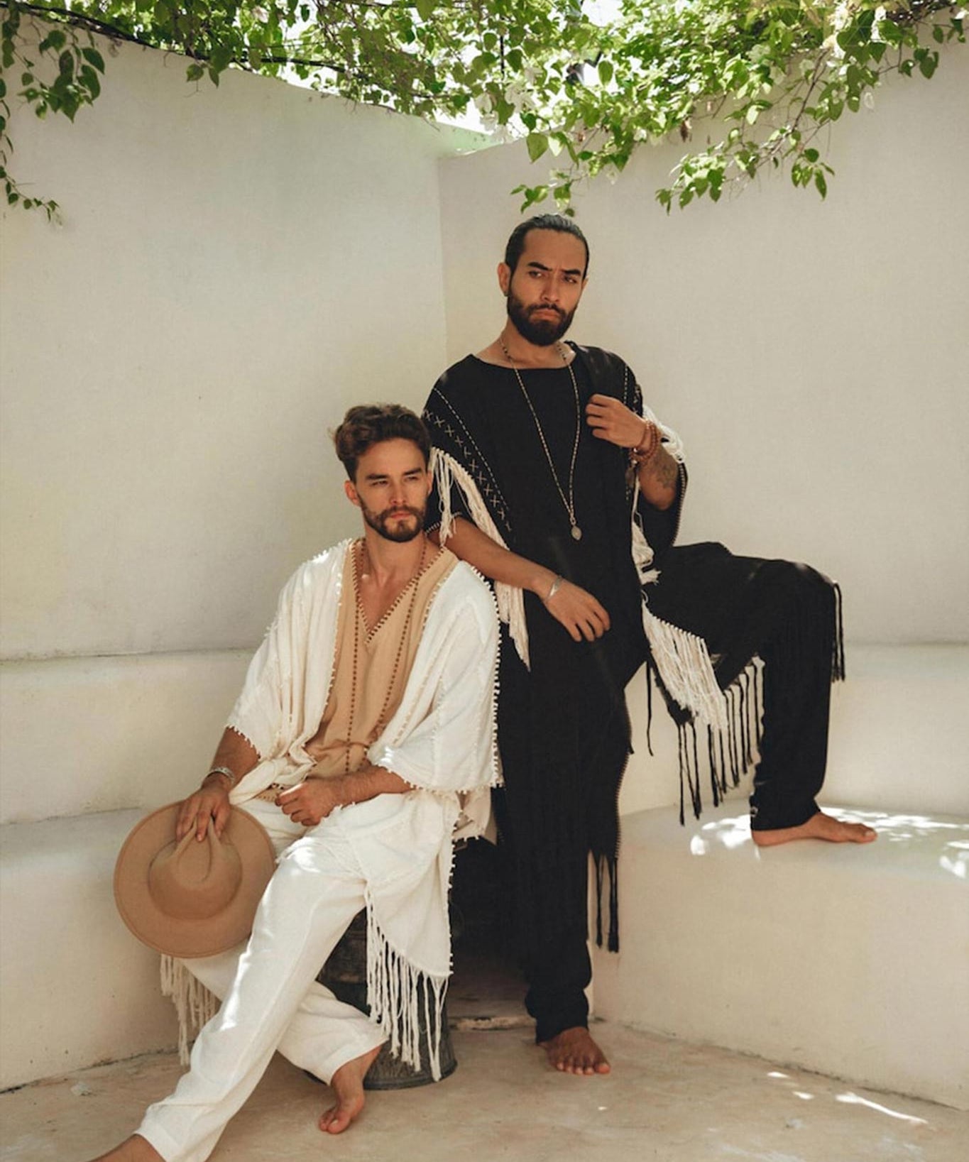 Two men in fringed outfits pose on a white bench under leafy branches. One sits, holding a hat, wearing the Off-White Long Cotton Pants by AYA Sacred Wear and an organic cotton shawl. The other stands confidently, dressed in black. The setting suggests a peaceful, outdoor environment.