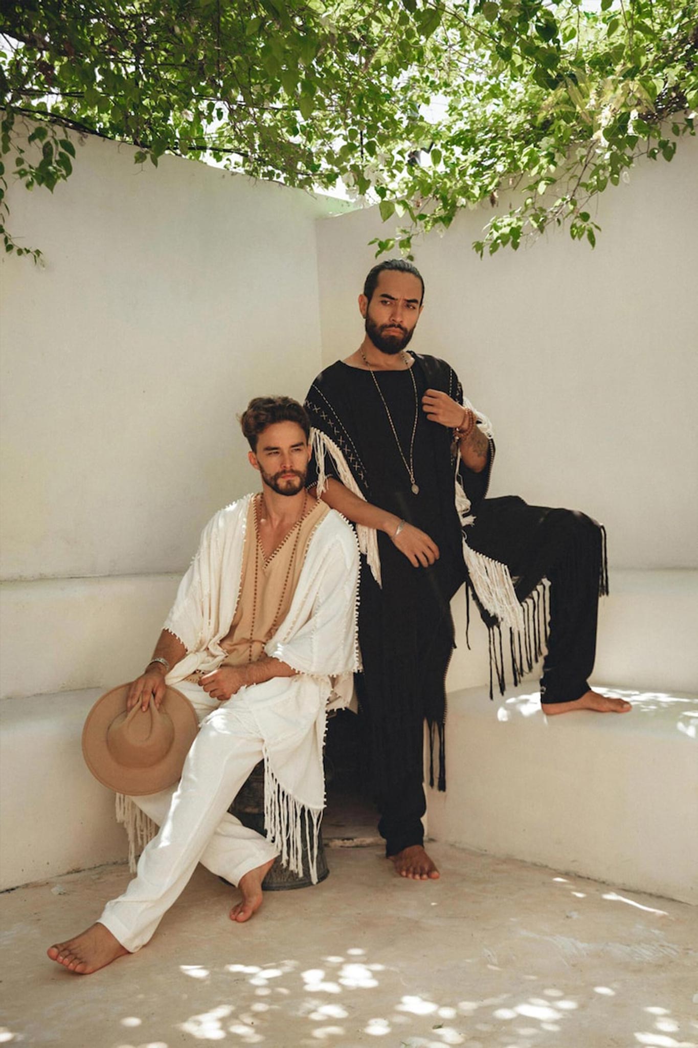Two men in fringed outfits pose on a white bench under leafy branches. One sits, holding a hat, wearing the Off-White Long Cotton Pants by AYA Sacred Wear and an organic cotton shawl. The other stands confidently, dressed in black. The setting suggests a peaceful, outdoor environment.
