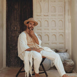 A man in a beige outfit and hat sits on a wooden chair in front of an ornate door. His ensemble includes AYA Sacred Wear's Off-White Long Cotton Pants for Men, blending seamlessly with earthy tones, clay pots, and intricate wall designs, creating a serene, rustic atmosphere.