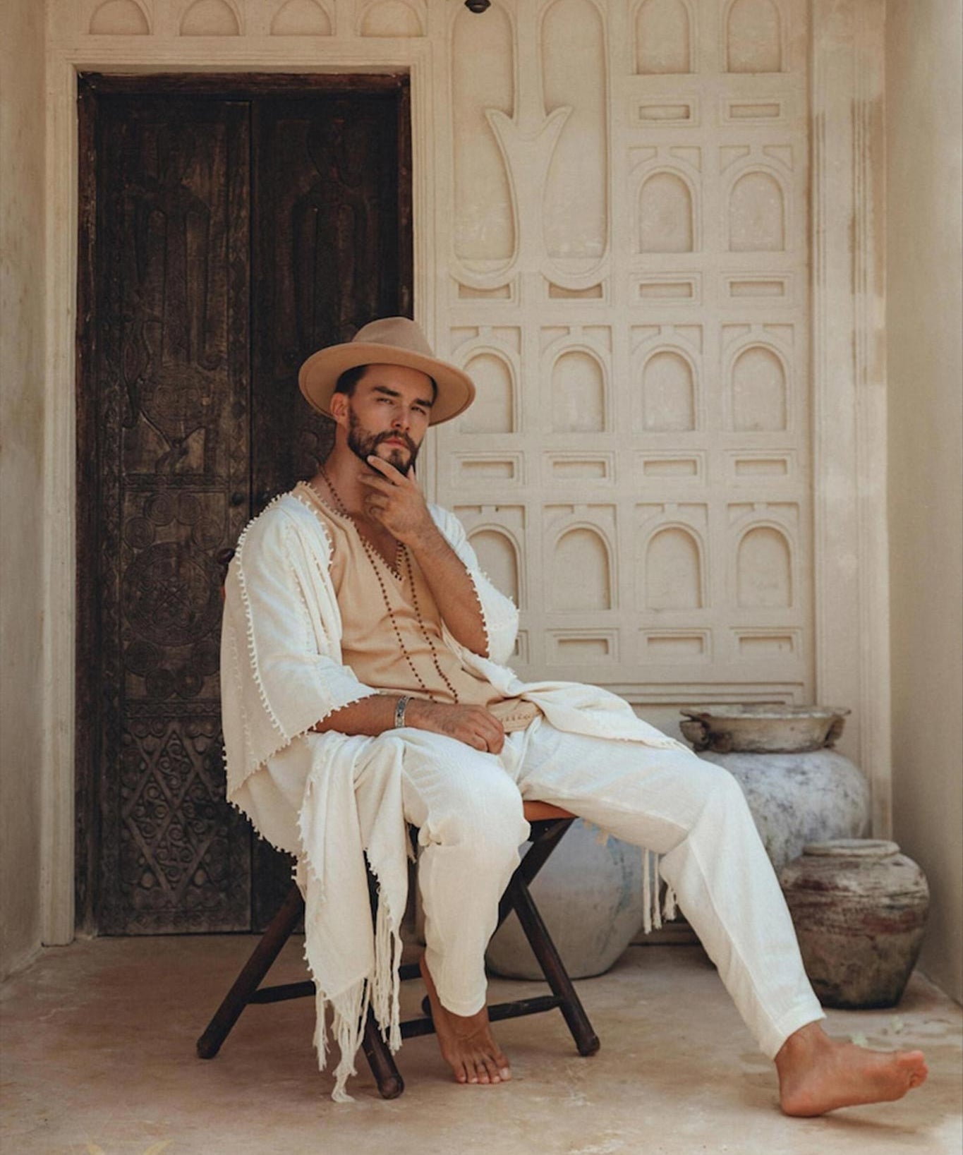 A man in a beige outfit and hat sits on a wooden chair in front of an ornate door. His ensemble includes AYA Sacred Wear's Off-White Long Cotton Pants for Men, blending seamlessly with earthy tones, clay pots, and intricate wall designs, creating a serene, rustic atmosphere.