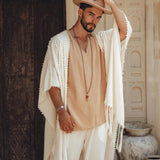 A man stands in front of an ornate wooden door wearing a beige shirt, a botanically dyed shawl, and a hat. His left hand rests on the brim of his hat as he accessorizes with a beaded necklace and Off-White Long Cotton Pants by AYA Sacred Wear. Two large clay pots are visible in the background.