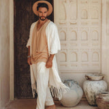 A man stands barefoot in a rustic setting, wearing a beige hat and an off-white, long fringed garment over a beige top and handwoven pants from AYA Sacred Wear's Off-White Long Cotton Pants for Men, crafted from organic cotton. He's surrounded by large, weathered clay pots and ornate architectural elements.