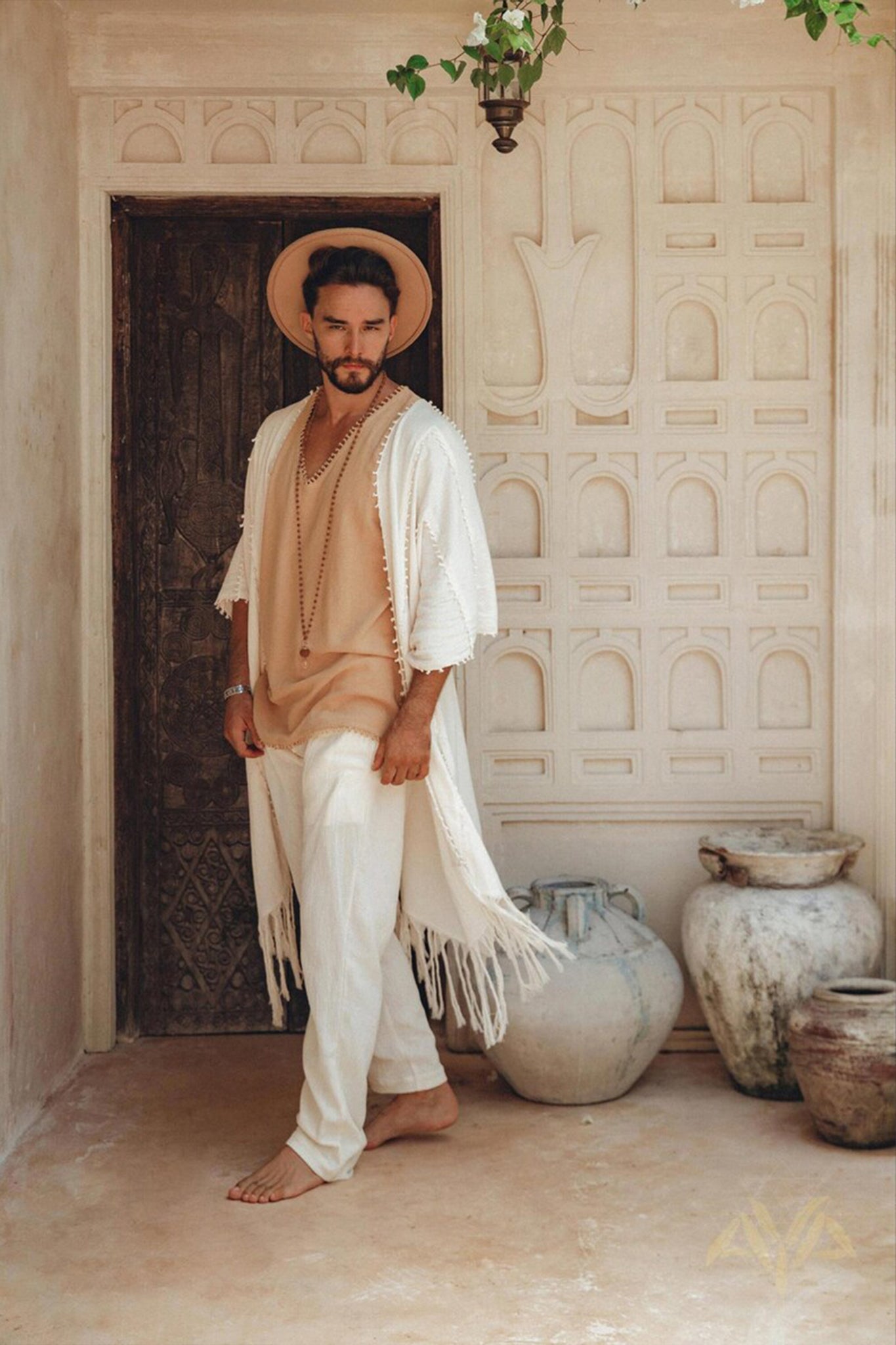 A man stands barefoot in a rustic setting, wearing a beige hat and an off-white, long fringed garment over a beige top and handwoven pants from AYA Sacred Wear's Off-White Long Cotton Pants for Men, crafted from organic cotton. He's surrounded by large, weathered clay pots and ornate architectural elements.