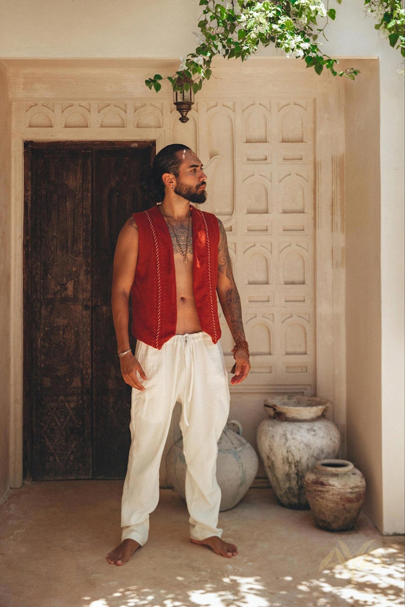 A bearded man with tattoos stands outdoors in front of an ornate wooden door, wearing an AYA Sacred Wear red vest and Off-White Long Cotton Pants for Men. Large clay pots are placed nearby, and a branch with green leaves hangs above him, showcasing the beauty of nature's handiwork.