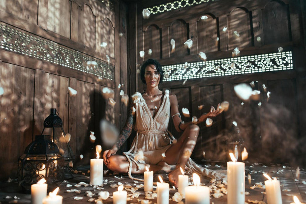 In a rustic wooden room filled with lit candles and scattered flower petals, a person sits cross-legged, wearing the Off-White Nomad Spirit Dress from AYA Sacred Wear. Crafted from organic fabric, this adjustable multiway dress enhances their relaxed demeanor as they gaze directly ahead. Ambient light casts intricate patterns on the walls.