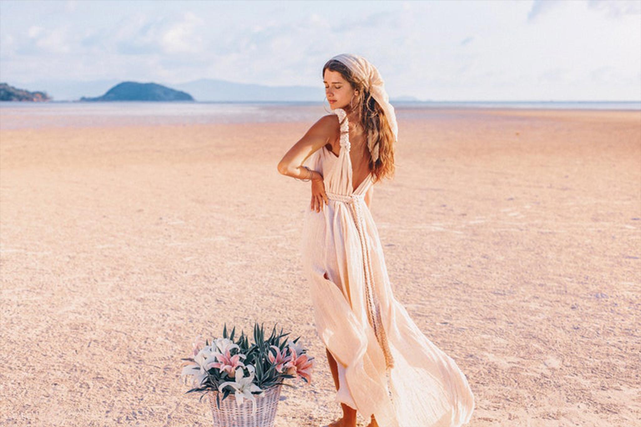 A woman in the Off-White Nomad Spirit Dress by AYA Sacred Wear, with a matching headscarf, stands on a tranquil sandy beach. She gazes down with a gentle expression beside a basket brimming with fresh pink flowers. In the background, the ocean and a distant island spread out under a clear sky.