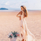 A woman in the Off-White Nomad Spirit Dress by AYA Sacred Wear, with a matching headscarf, stands on a tranquil sandy beach. She gazes down with a gentle expression beside a basket brimming with fresh pink flowers. In the background, the ocean and a distant island spread out under a clear sky.