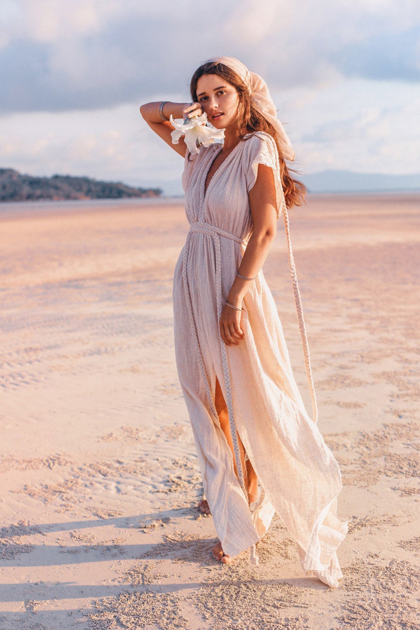 A woman in an Off-White Nomad Spirit Dress by AYA Sacred Wear, complemented by a headscarf, stands on a sunlit beach. She holds white flowers and gazes into the distance, with sand stretching to the horizon under a partly cloudy sky.