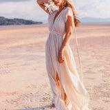A woman in an Off-White Nomad Spirit Dress by AYA Sacred Wear, complemented by a headscarf, stands on a sunlit beach. She holds white flowers and gazes into the distance, with sand stretching to the horizon under a partly cloudy sky.
