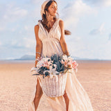 A woman in an AYA Sacred Wear Off-White Nomad Spirit Dress, elegantly flowing as she stands on a sandy beach with a large basket of pink and white flowers. The sky is clear with a few clouds, and the background showcases distant mountains, creating a serene, summery atmosphere.