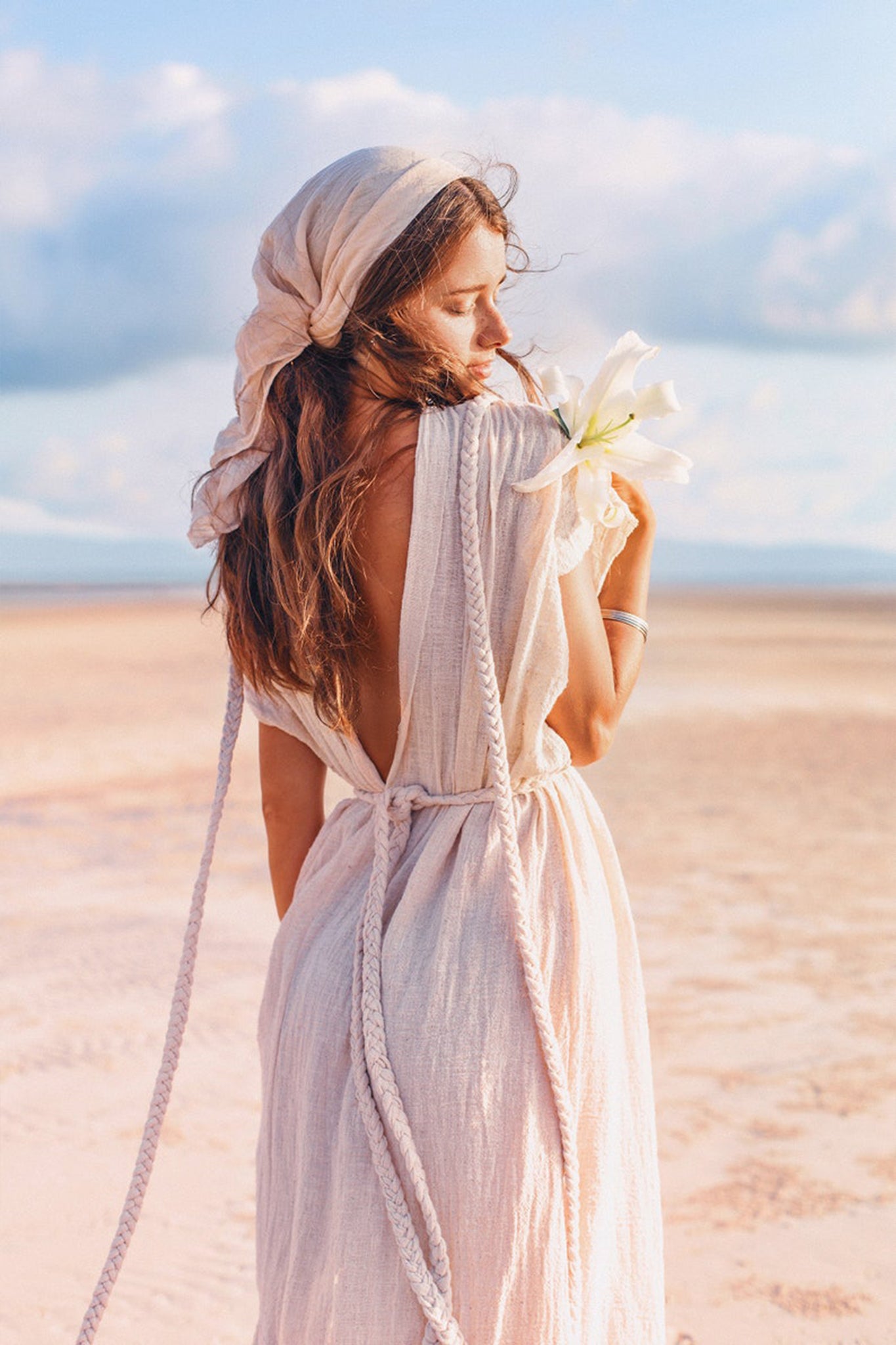 A woman dressed in an Off-White Nomad Spirit Dress from AYA Sacred Wear, featuring a backless, flowing design made of organic fabric and paired with a headscarf, stands on a sandy beach holding a white flower. The sky is blue with soft clouds as she glances over her shoulder, creating a serene and peaceful scene.