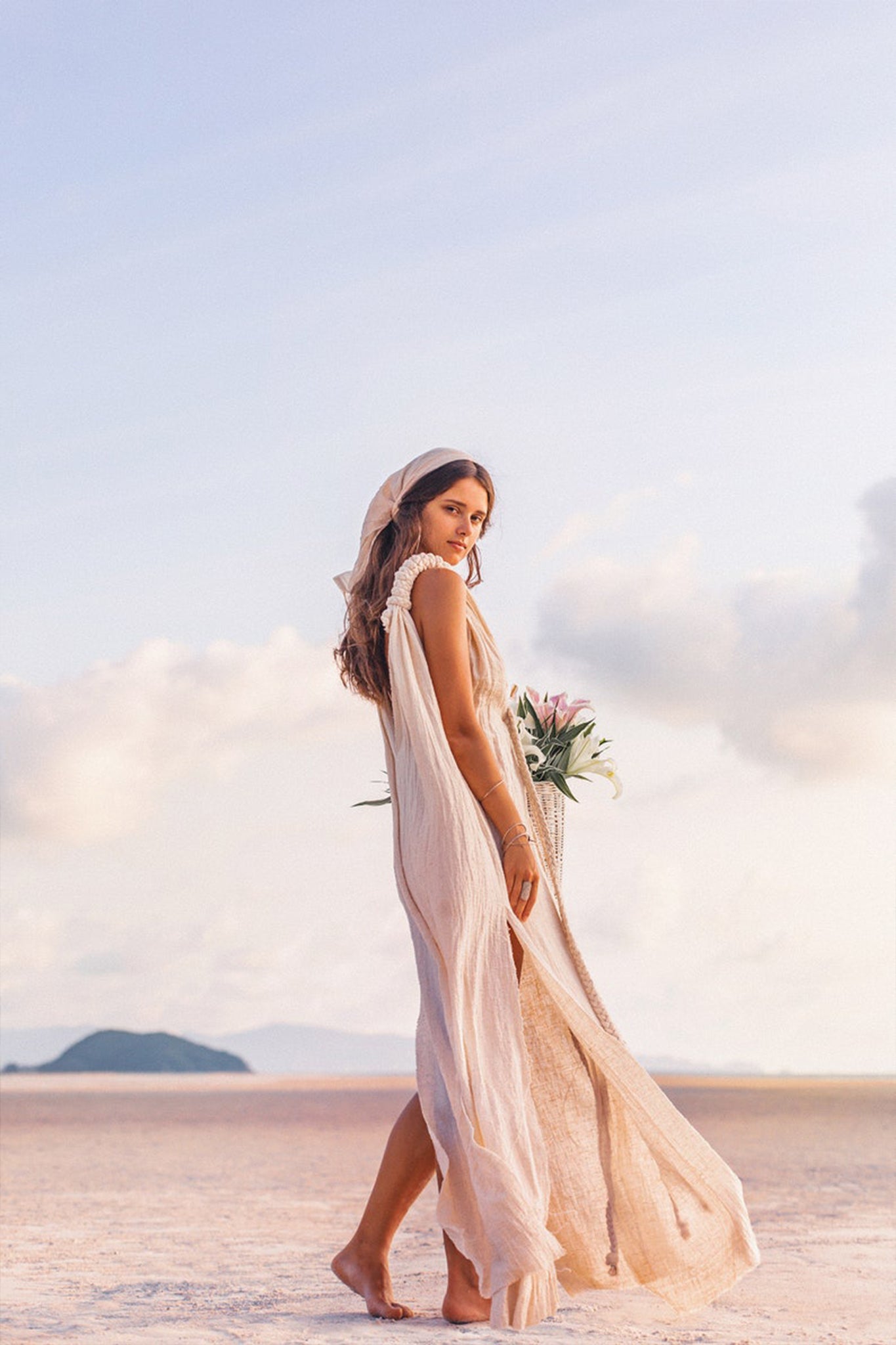 A woman wearing the Off-White Nomad Spirit Dress by AYA Sacred Wear, crafted from organic fabric, stands gracefully on a sandy beach with a bouquet of flowers in her hands. The clear sky shows a few clouds as an island appears in the distance. She glances over her shoulder with a gentle smile.