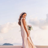 A woman wearing the Off-White Nomad Spirit Dress by AYA Sacred Wear, crafted from organic fabric, stands gracefully on a sandy beach with a bouquet of flowers in her hands. The clear sky shows a few clouds as an island appears in the distance. She glances over her shoulder with a gentle smile.