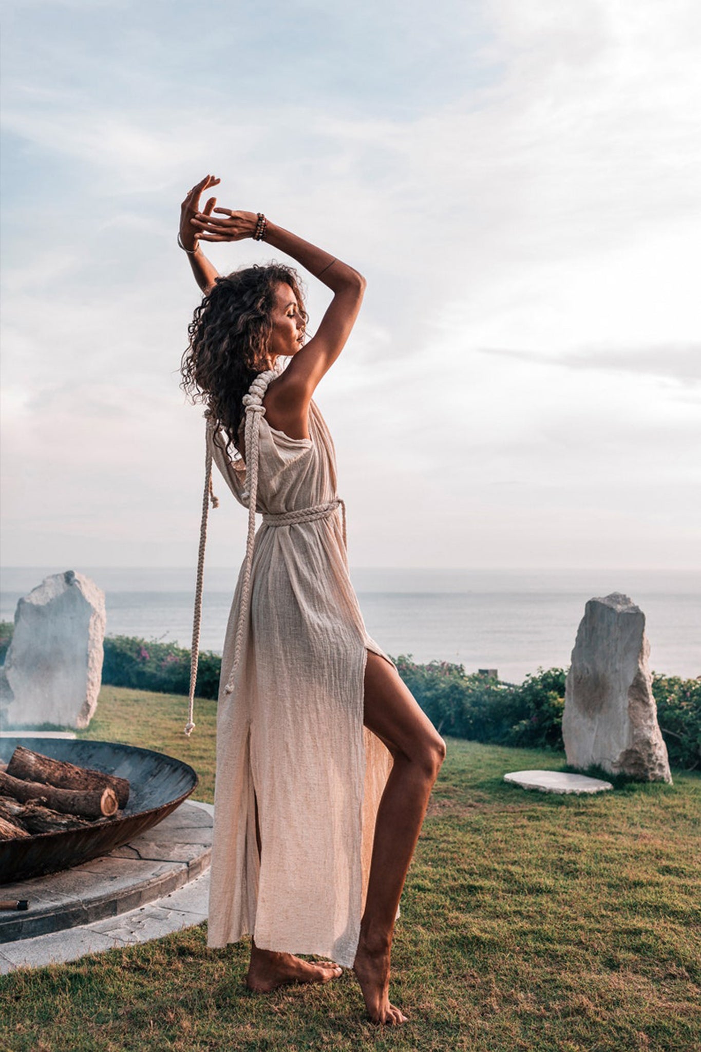 A person with curly hair poses gracefully outdoors by a large fire pit. They are wearing the Off-White Nomad Spirit Dress by AYA Sacred Wear, a light, sleeveless boho wedding dress featuring a high slit. Standing barefoot on grass near a coastline under a partly cloudy sky, they exude an elegant natural beauty.