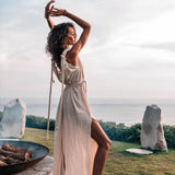 A person with curly hair poses gracefully outdoors by a large fire pit. They are wearing the Off-White Nomad Spirit Dress by AYA Sacred Wear, a light, sleeveless boho wedding dress featuring a high slit. Standing barefoot on grass near a coastline under a partly cloudy sky, they exude an elegant natural beauty.
